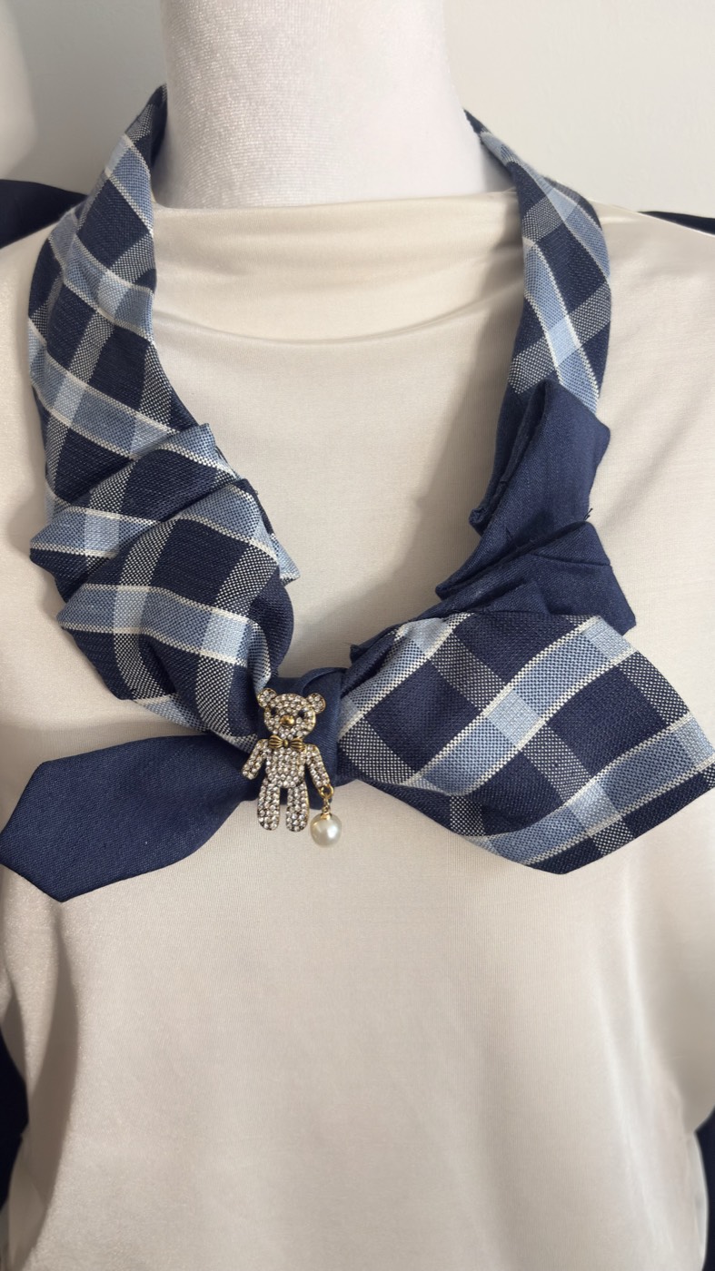 Navy and light blue plaid necktie tied into a soft scarf-style knot with a crystal teddy bear brooch and a pearl drop.